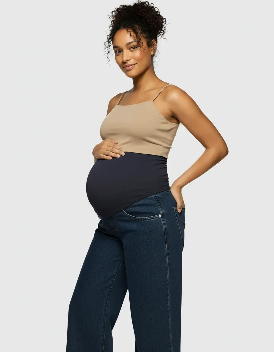 Smiling pregnant woman in a beige camisole and dark wash wide-leg maternity jeans with a black elastic waist.