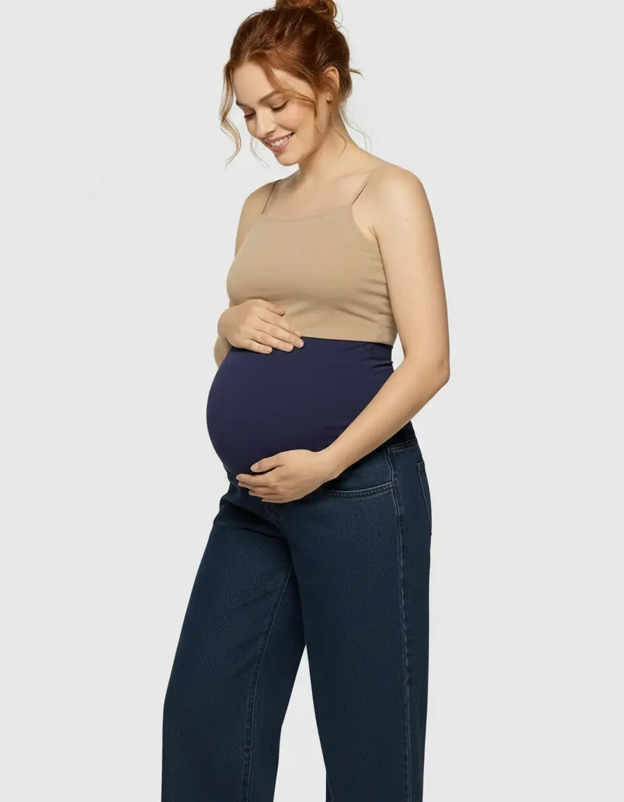 Smiling pregnant woman in dark wash wide-leg maternity jeans with a navy elastic waist and a beige tank top.