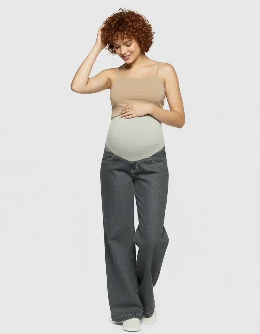 Smiling pregnant woman in dark grey wide-leg maternity jeans with a light grey elastic waist and a beige tank top.