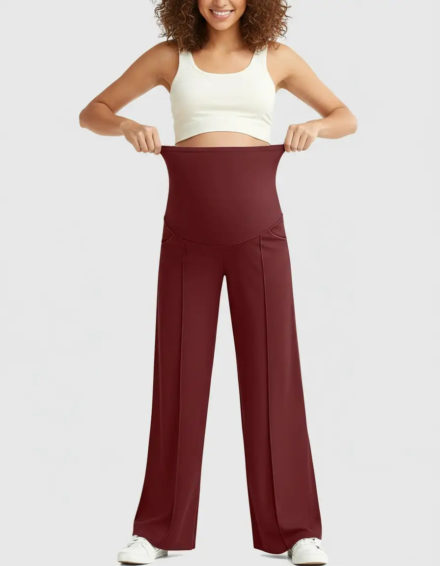 Smiling woman in a white crop top holding up the high waist of relaxed-fit burgundy maternity pants.