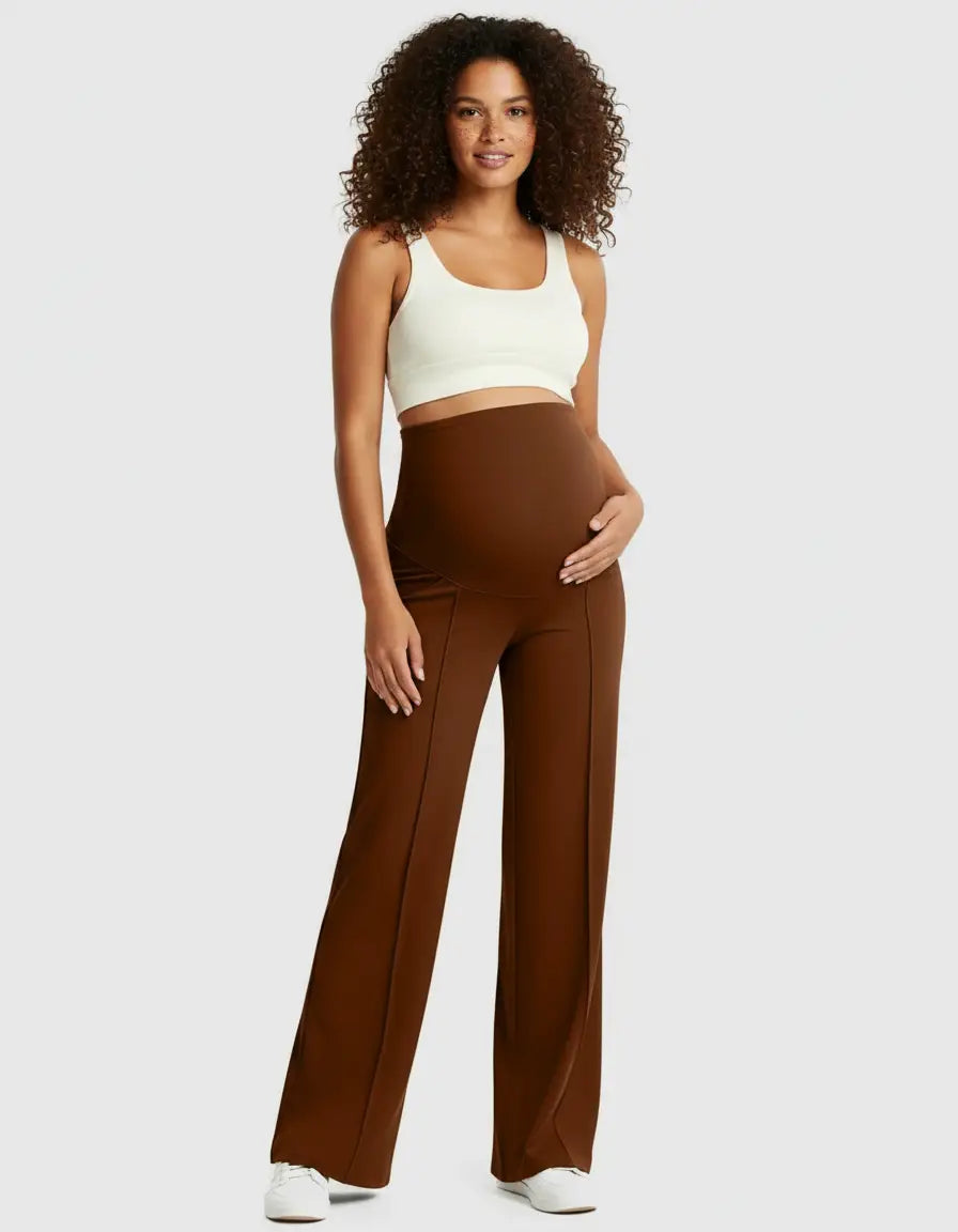Smiling pregnant woman in brown high-waist maternity pants and a white crop top, cradling her bump.