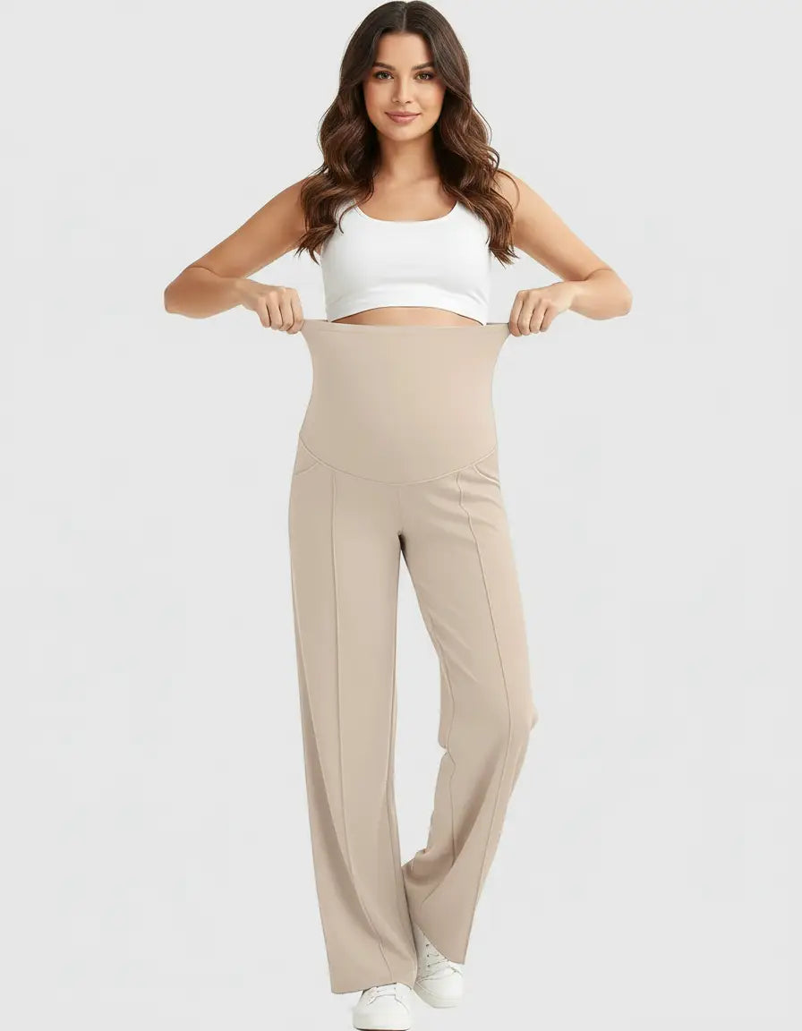Smiling pregnant woman in a white crop top holding up beige high-waist maternity pants with a relaxed fit.