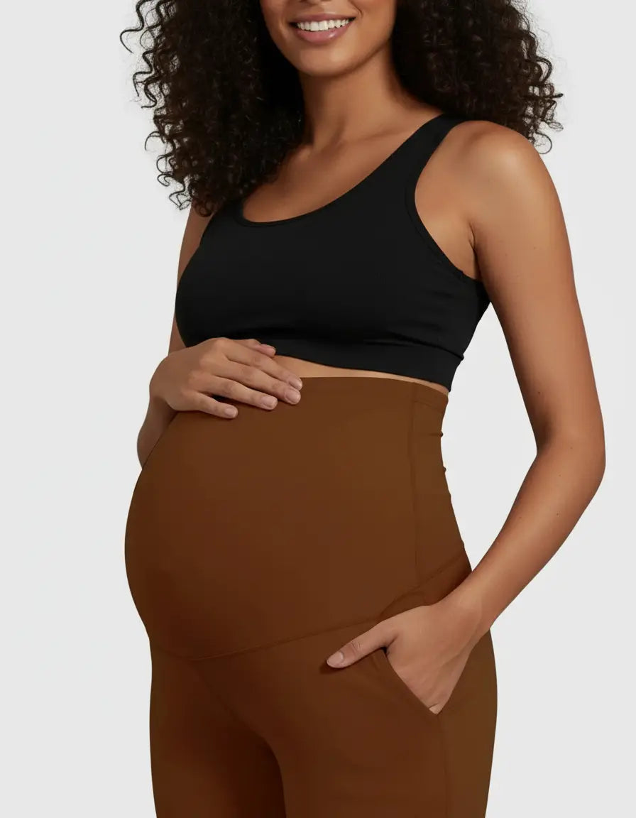 Smiling pregnant woman in a black sports bra and brown high-waist straight-leg maternity pants with pockets.