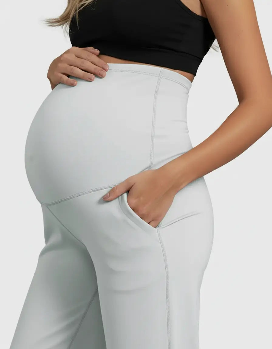 Pregnant woman in light gray high-waist straight-leg maternity pants with pockets, holding her baby bump.