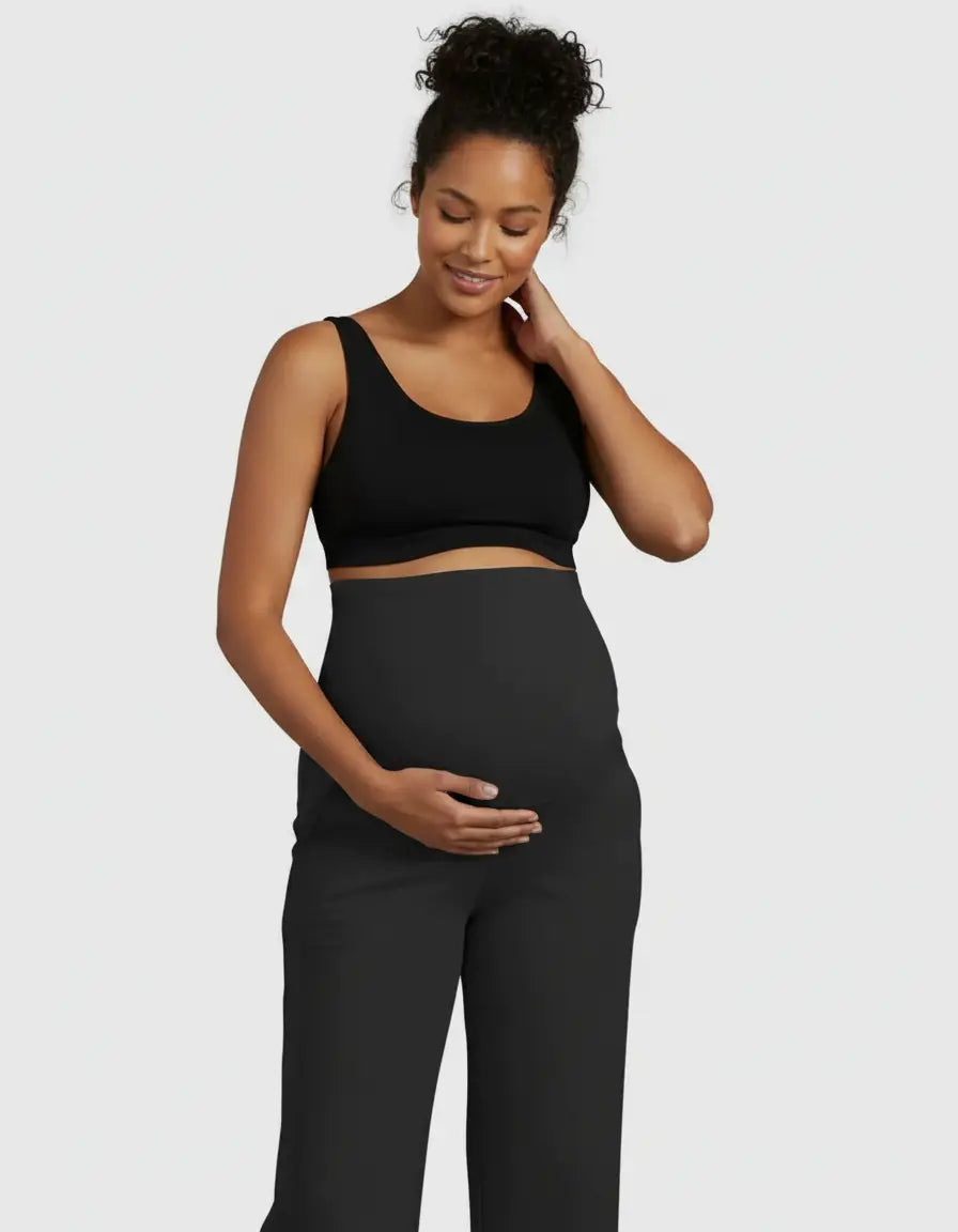 Smiling pregnant woman in black high-waist straight-leg maternity pants and a black bra top, cradling her bump.