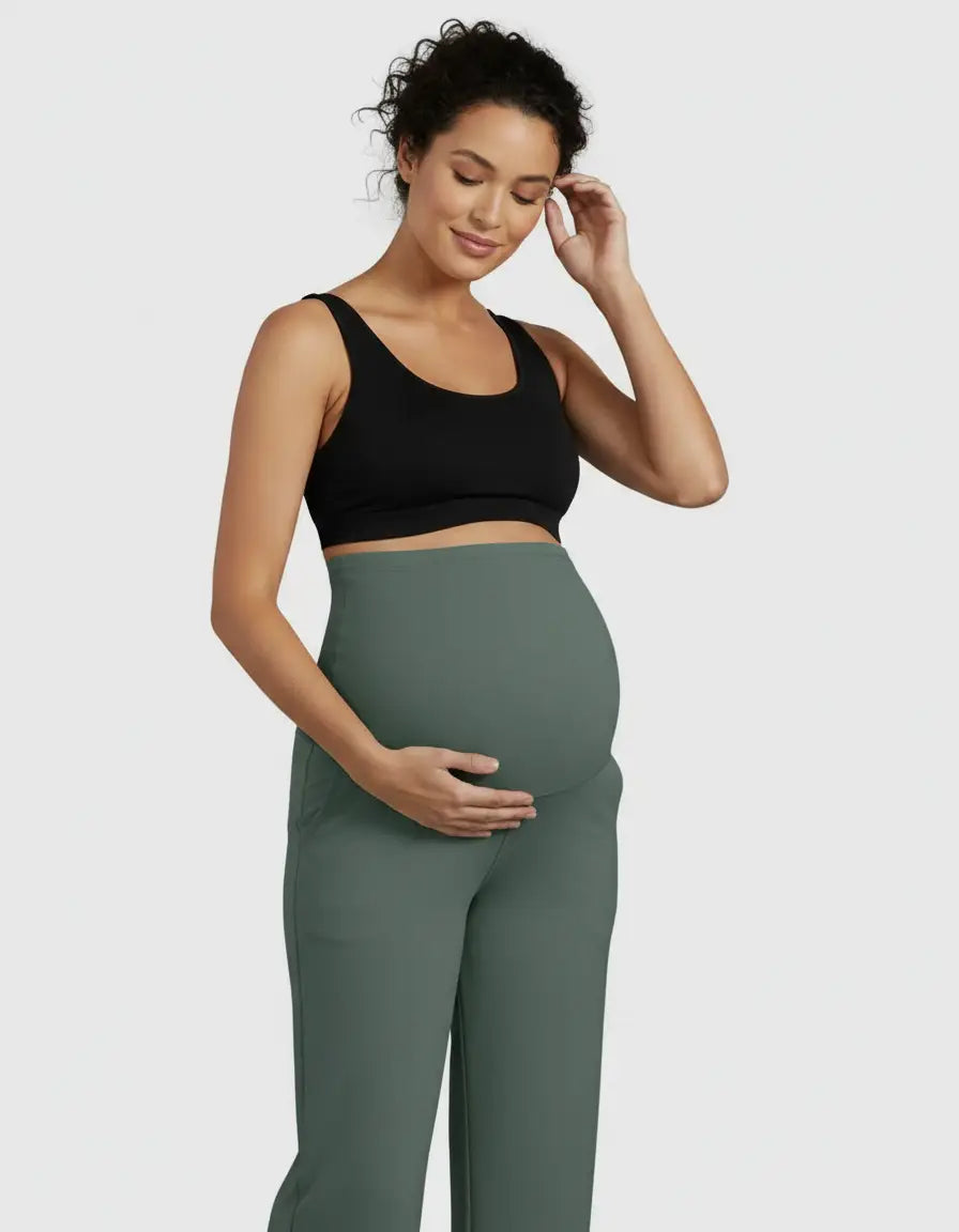 Pregnant woman in a black bra and sage green high-waist straight-leg maternity pants, gently touching her belly.