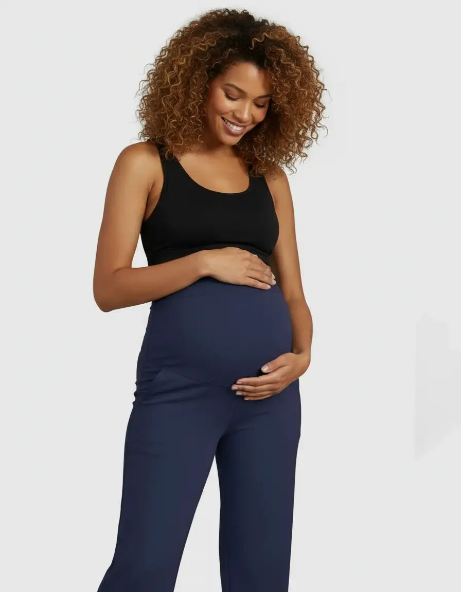 Smiling pregnant woman in a black top and navy high-waist straight-leg maternity pants, cradling her bump.