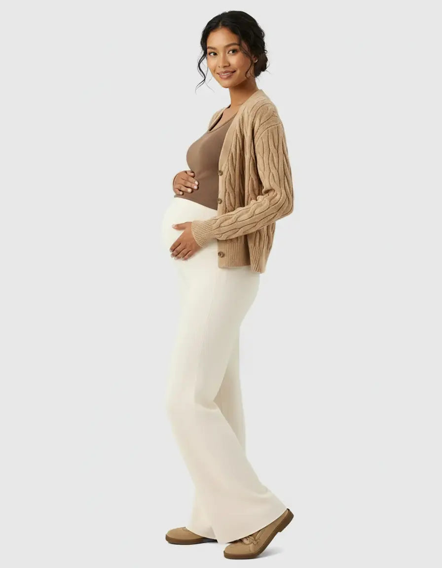 Smiling pregnant woman in a brown top, cable-knit cardigan, and cream wide-leg maternity wool pants.