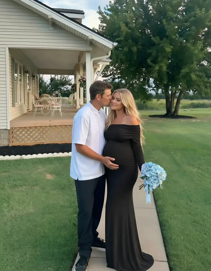 Pregnant woman in a black off-shoulder cross-back maternity gown with partner, holding blue flowers.