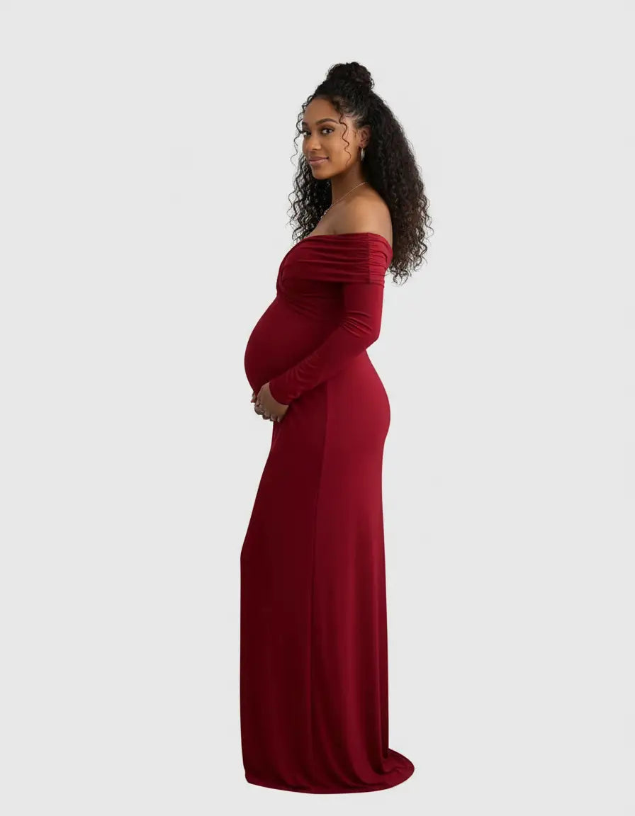 Pregnant woman in a long, red off-shoulder cross-back maternity gown, gently cradling her baby bump.