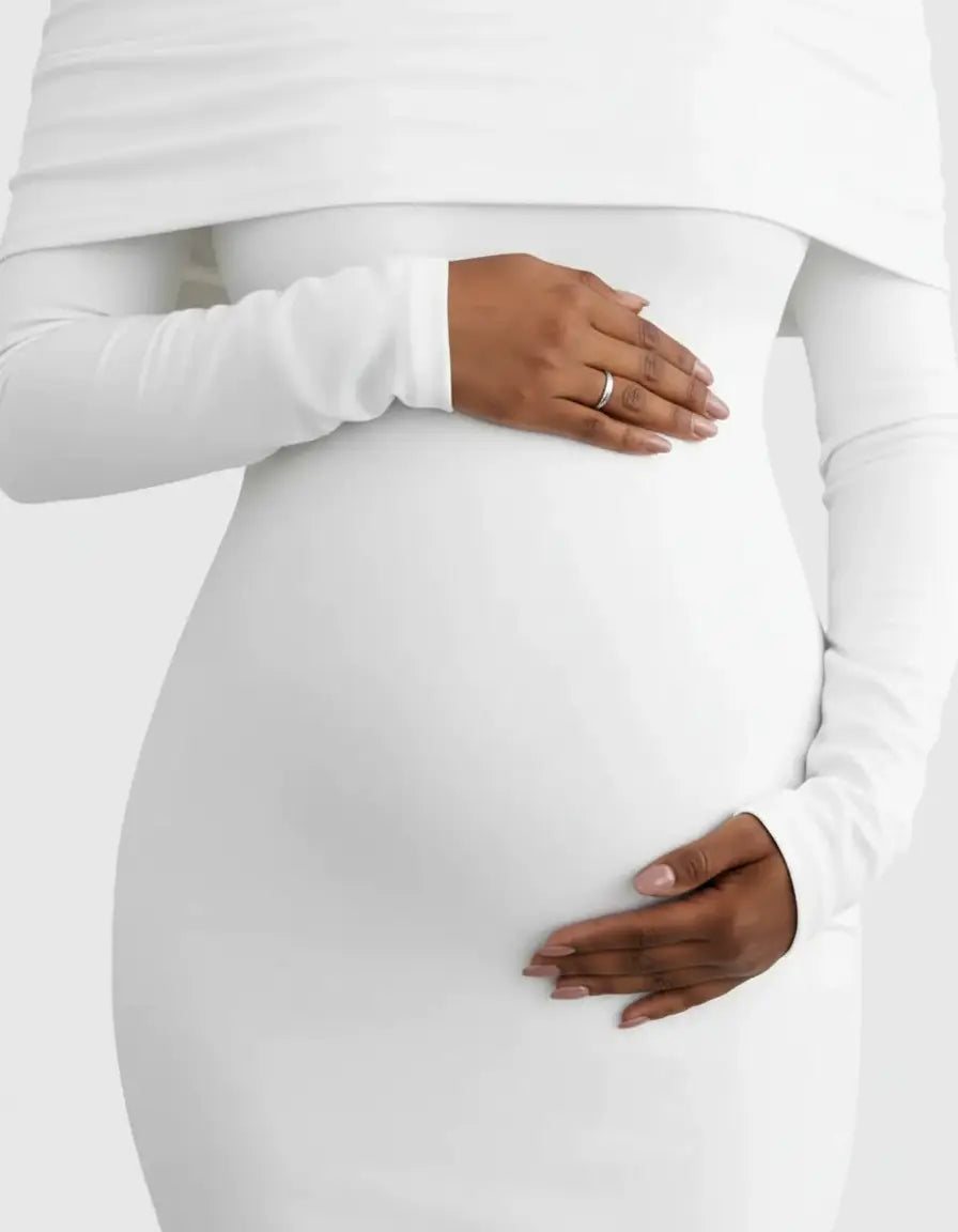 Pregnant woman in a white off-shoulder cross-back maternity gown, hands gently on her baby bump.