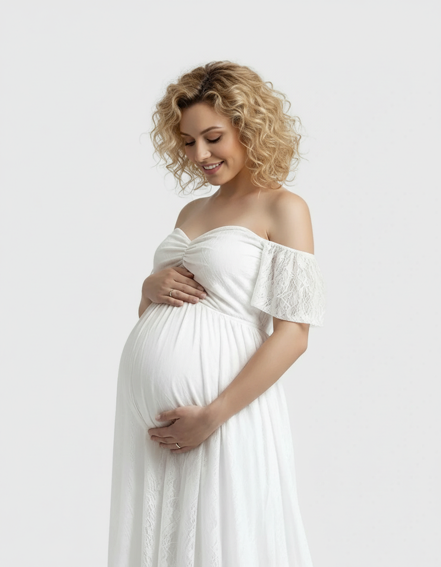 Smiling pregnant woman in a white off-shoulder flowing lace maternity maxi dress, cradling her baby bump.
