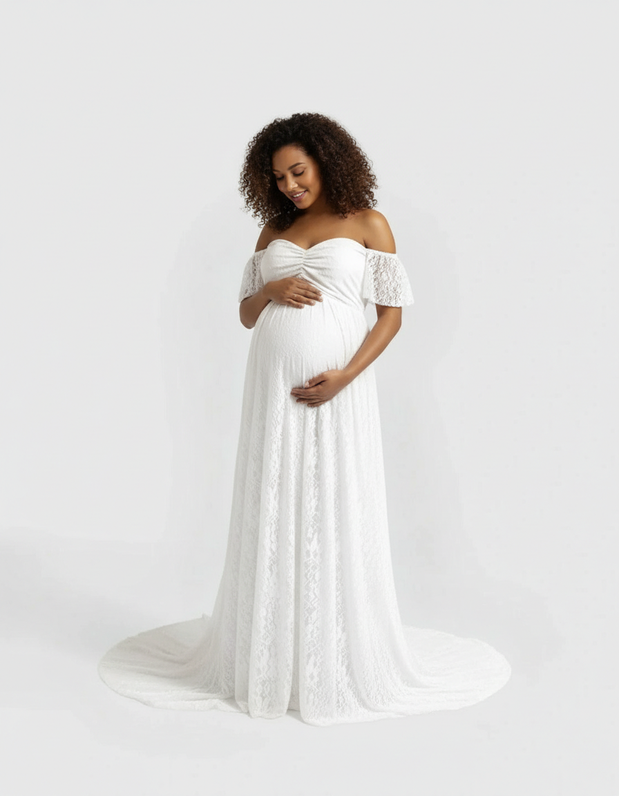 Smiling pregnant woman in a white off-shoulder flowing lace maternity maxi dress, hands on belly.