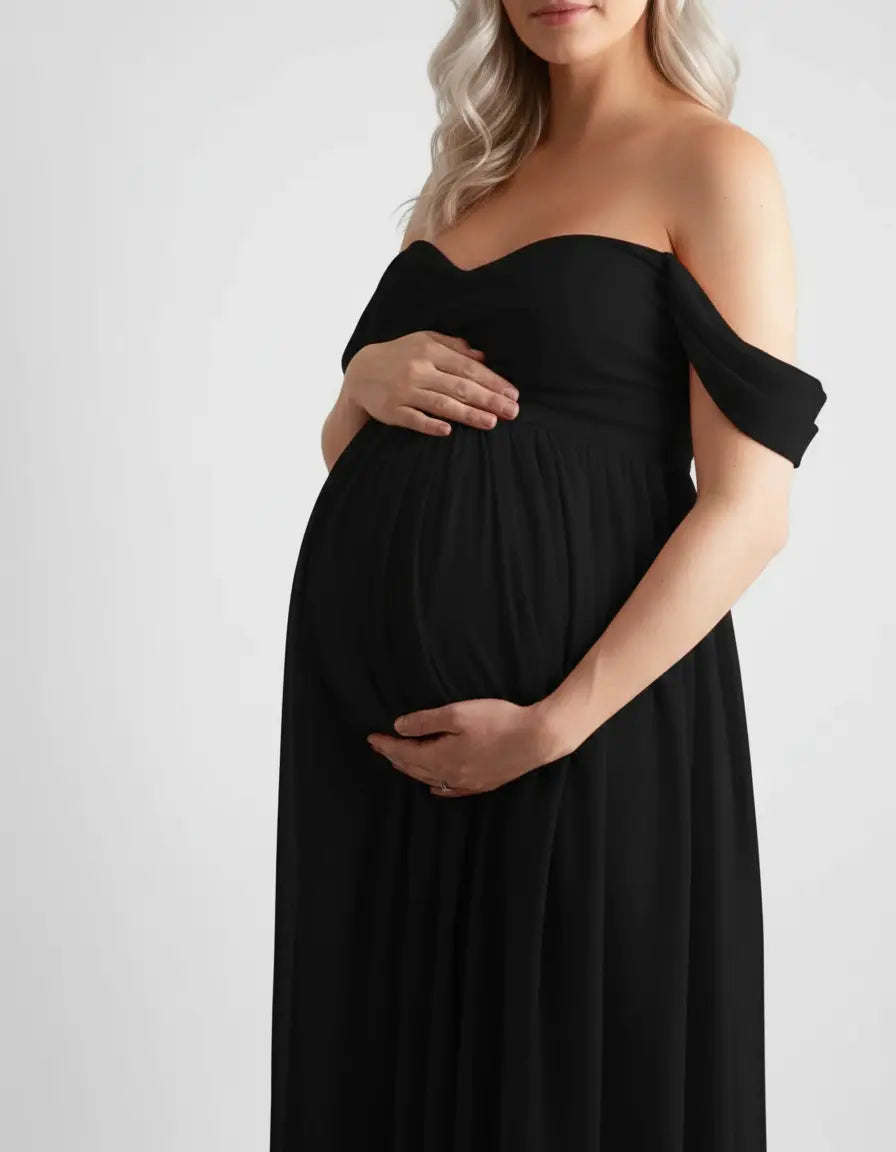 Pregnant woman in a black off-shoulder flowing maternity photoshoot gown, cradling her baby bump.