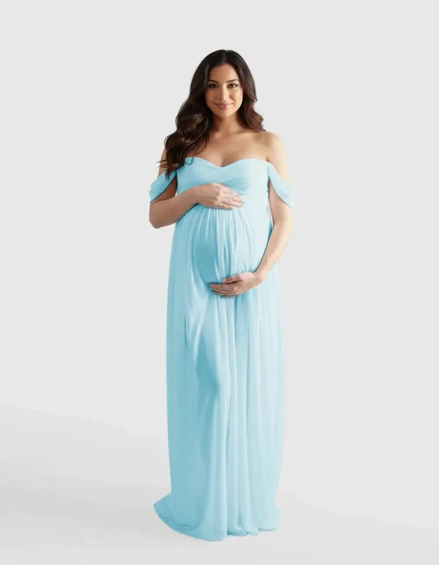 Pregnant woman in a flowing, light blue off-shoulder maternity photoshoot gown, hands on belly.