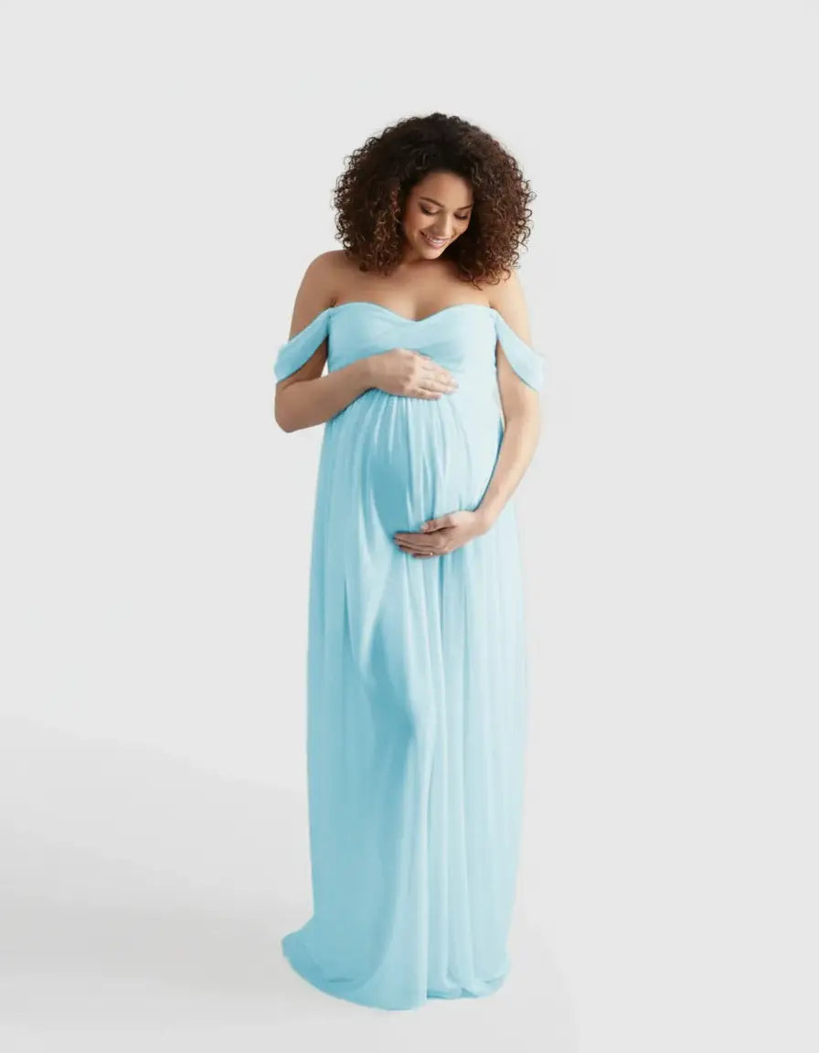 Smiling pregnant woman in a light blue off-shoulder flowing maternity photoshoot gown, cradling her bump.