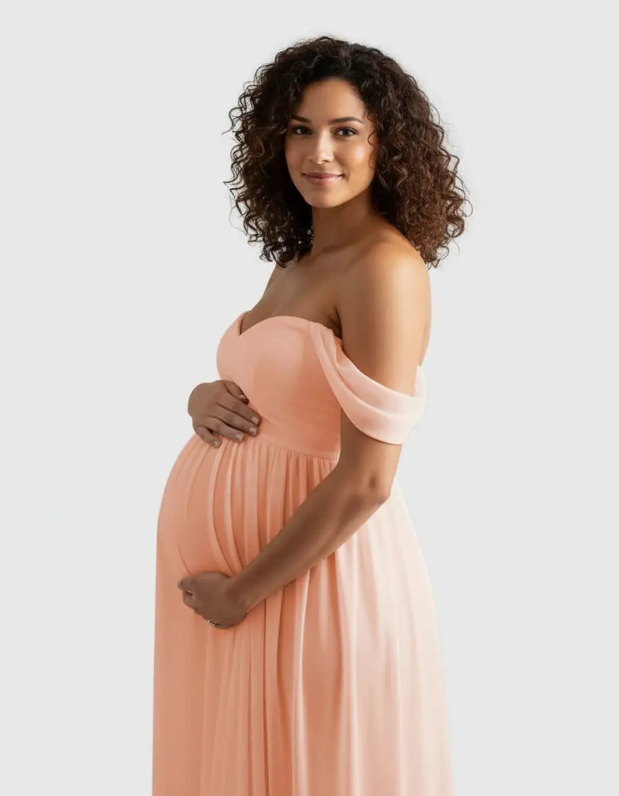 Pregnant woman in a flowing peach off-shoulder maternity photoshoot gown, gently cradling her bump.