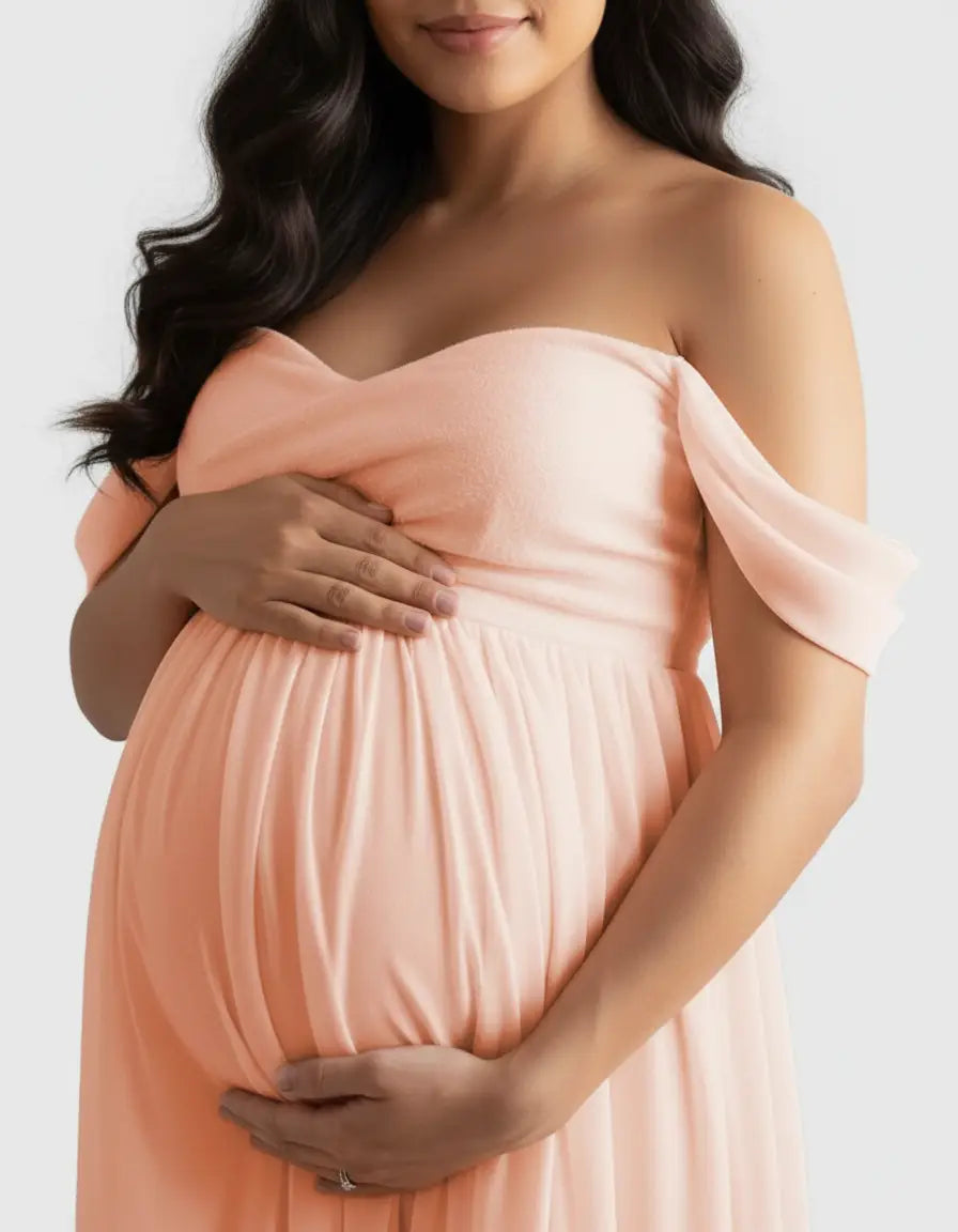 Pregnant woman in a flowing, peach off-shoulder maternity photoshoot gown, cradling her baby bump.