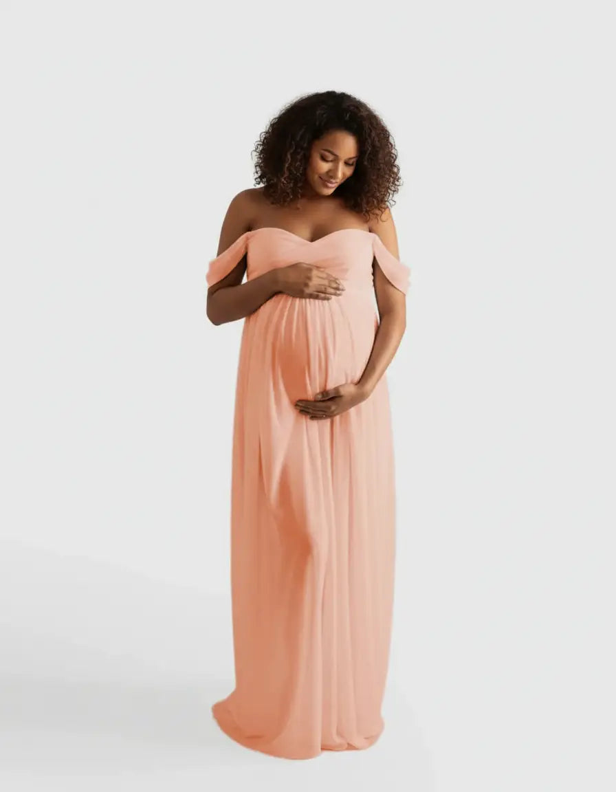 Pregnant woman in a flowing, off-shoulder peach maternity photoshoot gown, gently cradling her belly.