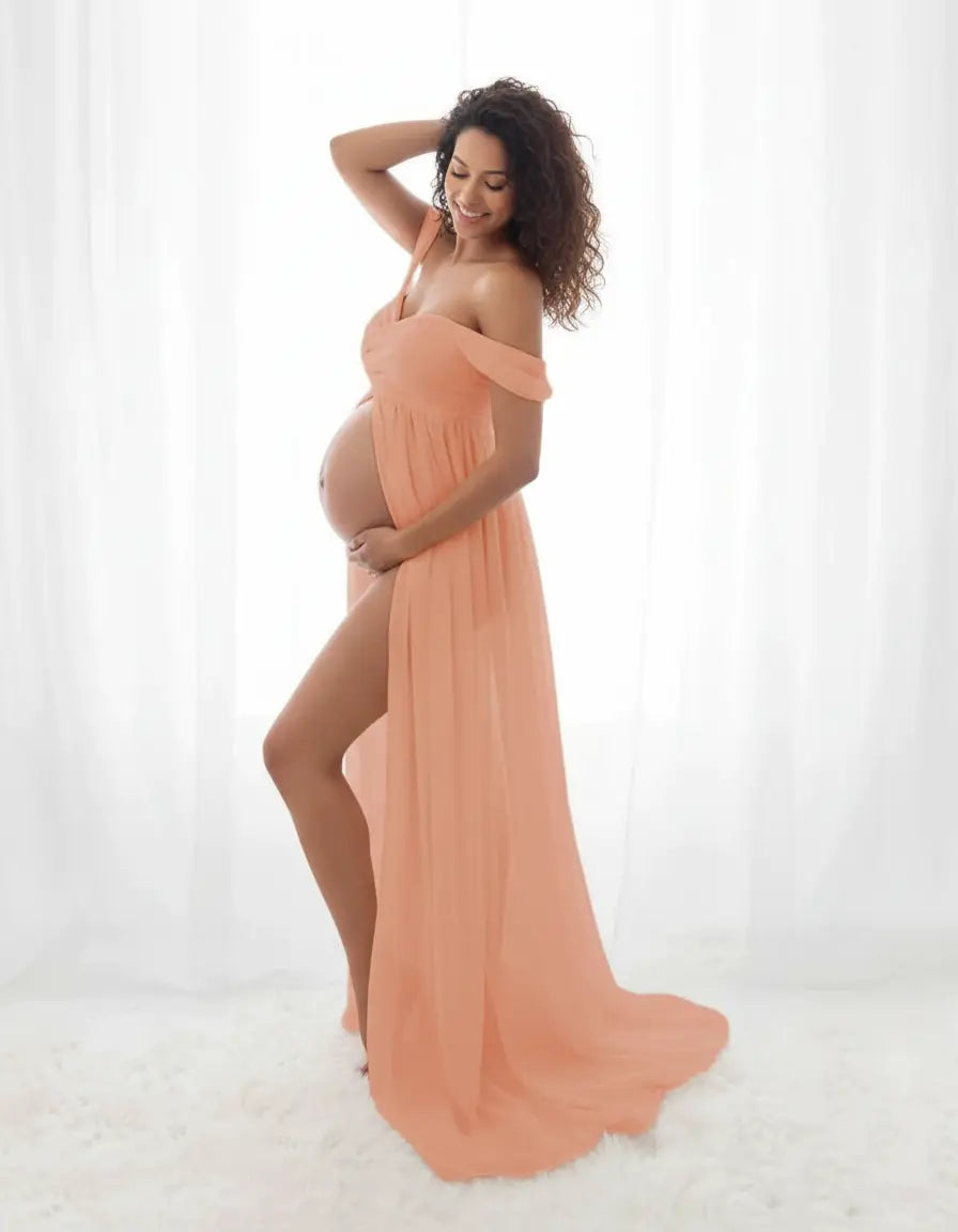 Smiling pregnant woman in a flowing, off-shoulder peach maternity photoshoot gown with a high slit.