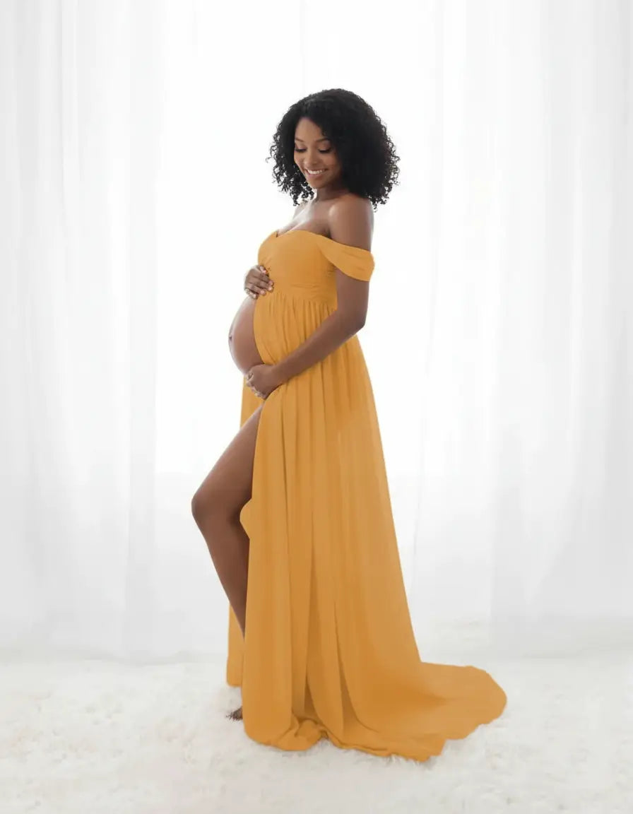 Smiling pregnant woman in a flowing mustard yellow off-shoulder maternity photoshoot gown with a thigh-high slit.