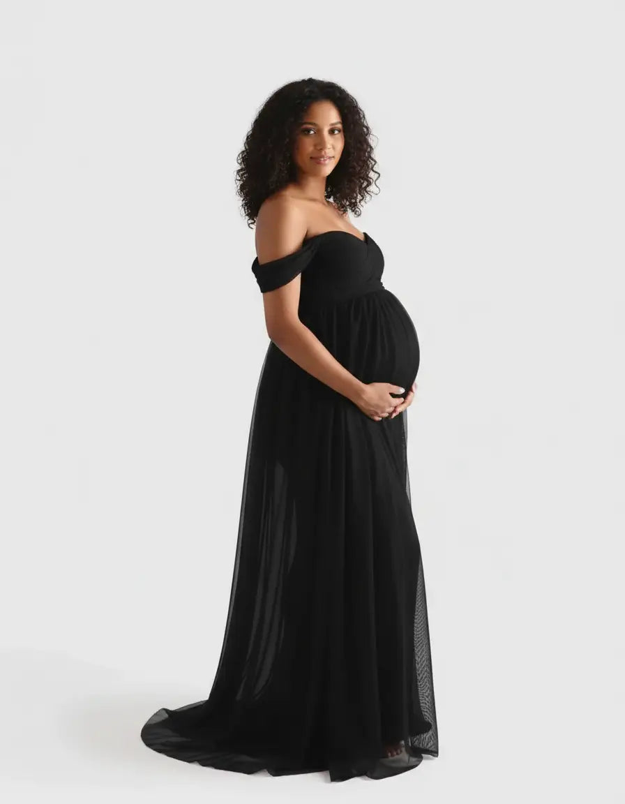 Pregnant woman in a black off-shoulder flowing maternity photoshoot gown, cradling her baby bump.