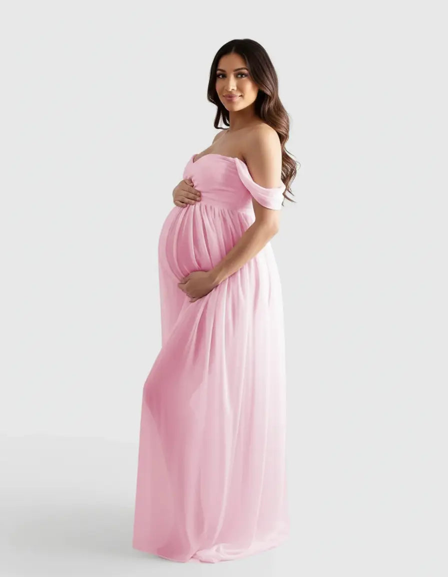 Smiling pregnant woman in a flowing, off-shoulder pink maternity photoshoot gown, cradling her baby bump.