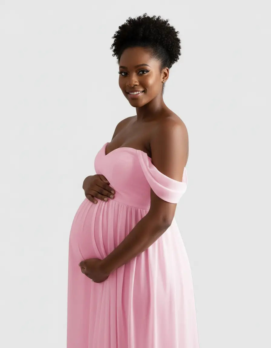 Smiling pregnant woman in a flowing pink off-shoulder maternity photoshoot gown, cradling her bump.