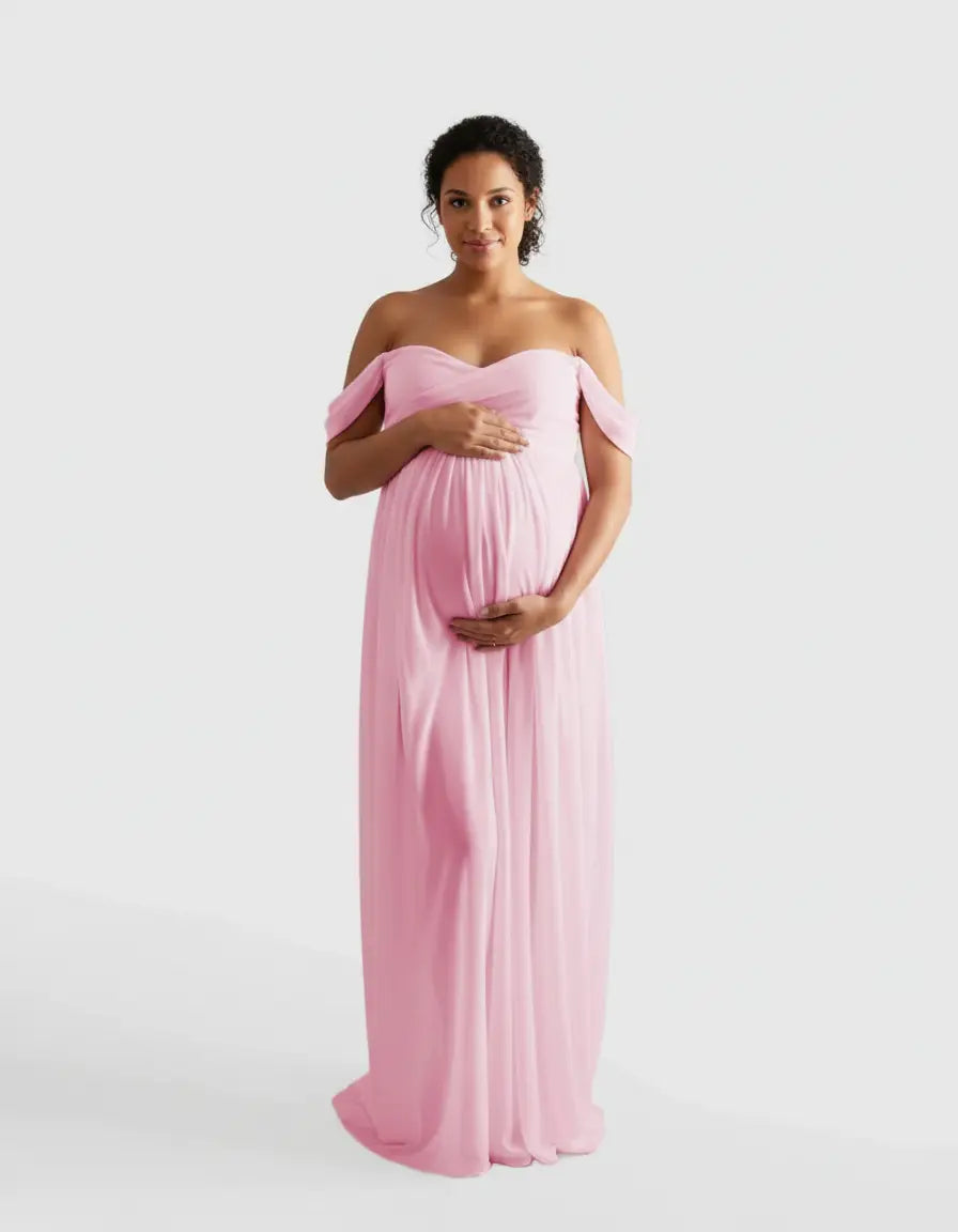 Maternity model in a pink off-shoulder flowing gown, cradling her baby bump.