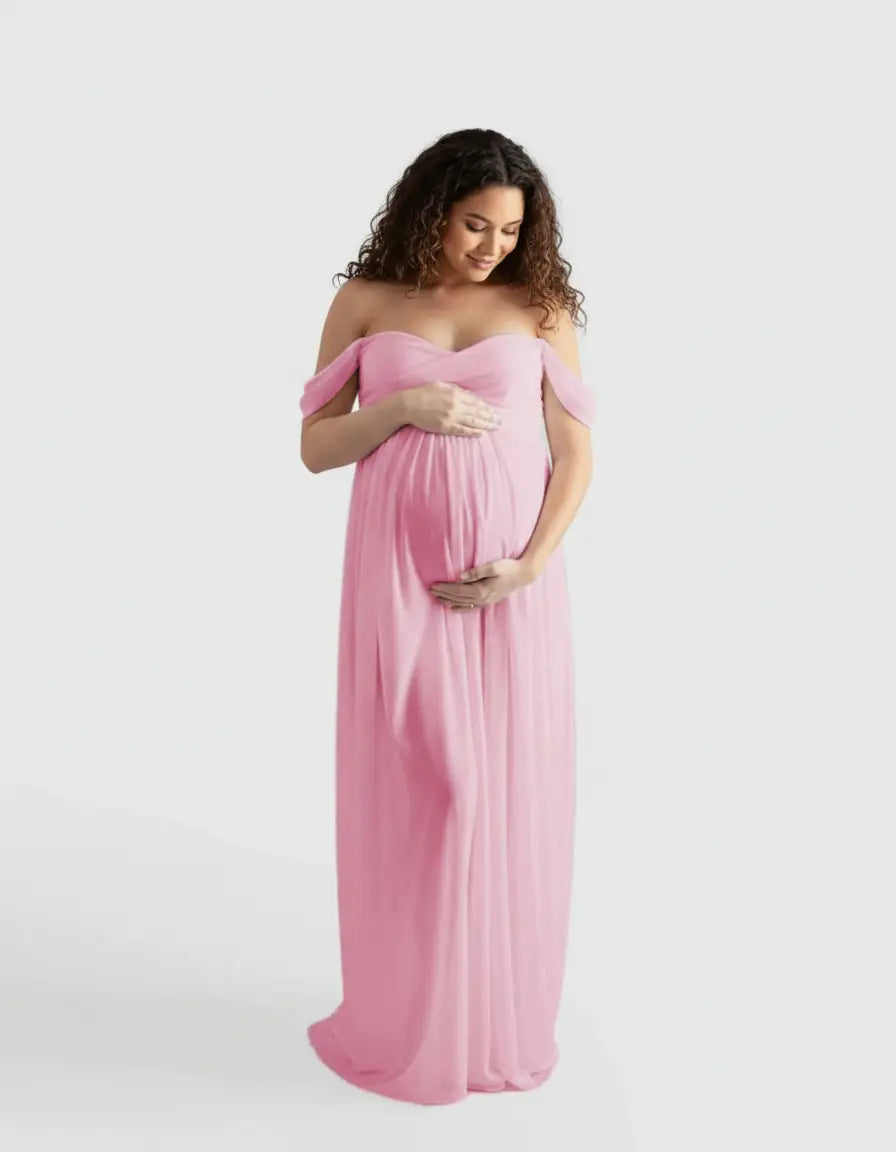 Pregnant woman in a pink off-shoulder flowing maternity photoshoot gown, gently cradling her belly.