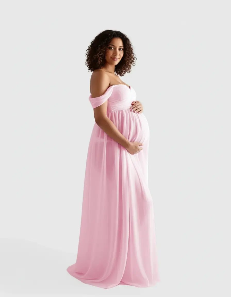 Smiling pregnant woman in a flowing, light pink off-shoulder maternity photoshoot gown.