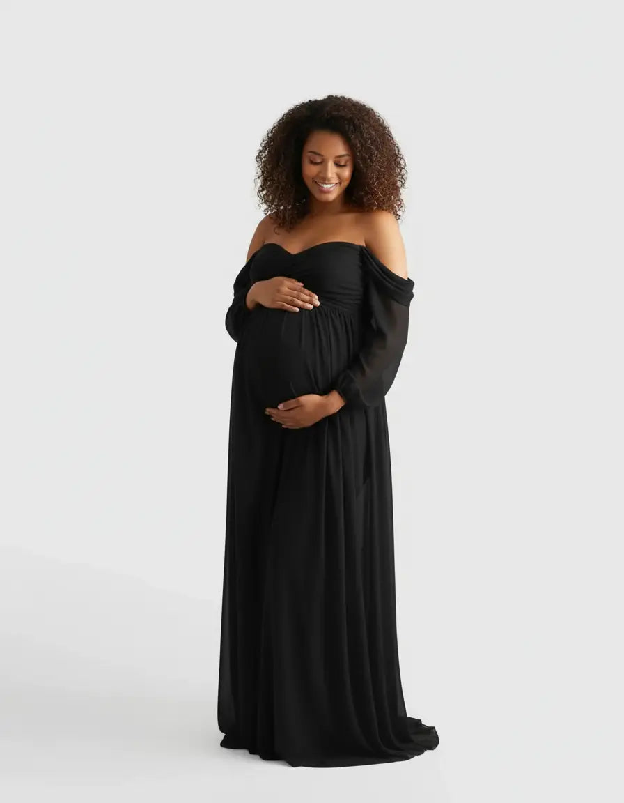 Smiling pregnant woman in a black off-shoulder flowing maternity photoshoot gown, cradling her baby bump.
