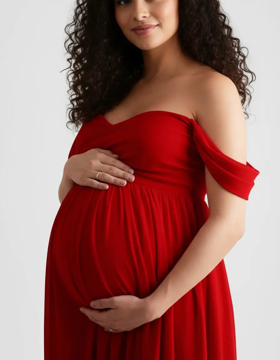 Pregnant woman in a flowing red off-shoulder maternity photoshoot gown, cradling her baby bump.