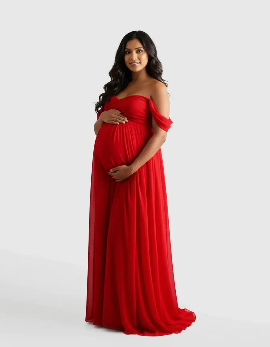 Pregnant woman in a flowing red off-shoulder maternity photoshoot gown, holding her baby bump.