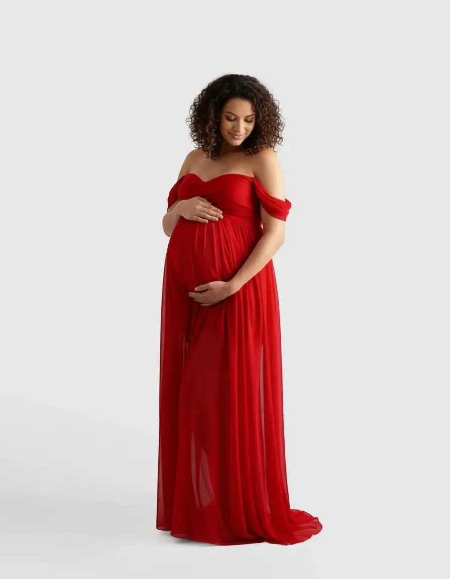Pregnant woman in a flowing red off-shoulder maternity photoshoot gown, gently cradling her bump.