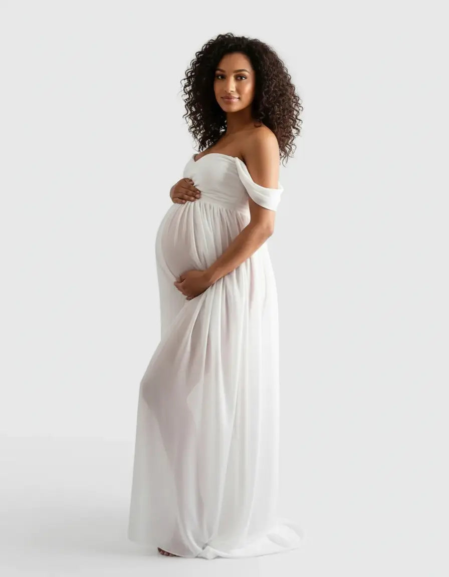 Pregnant woman in a white off-shoulder flowing maternity photoshoot gown, cradling her baby bump.
