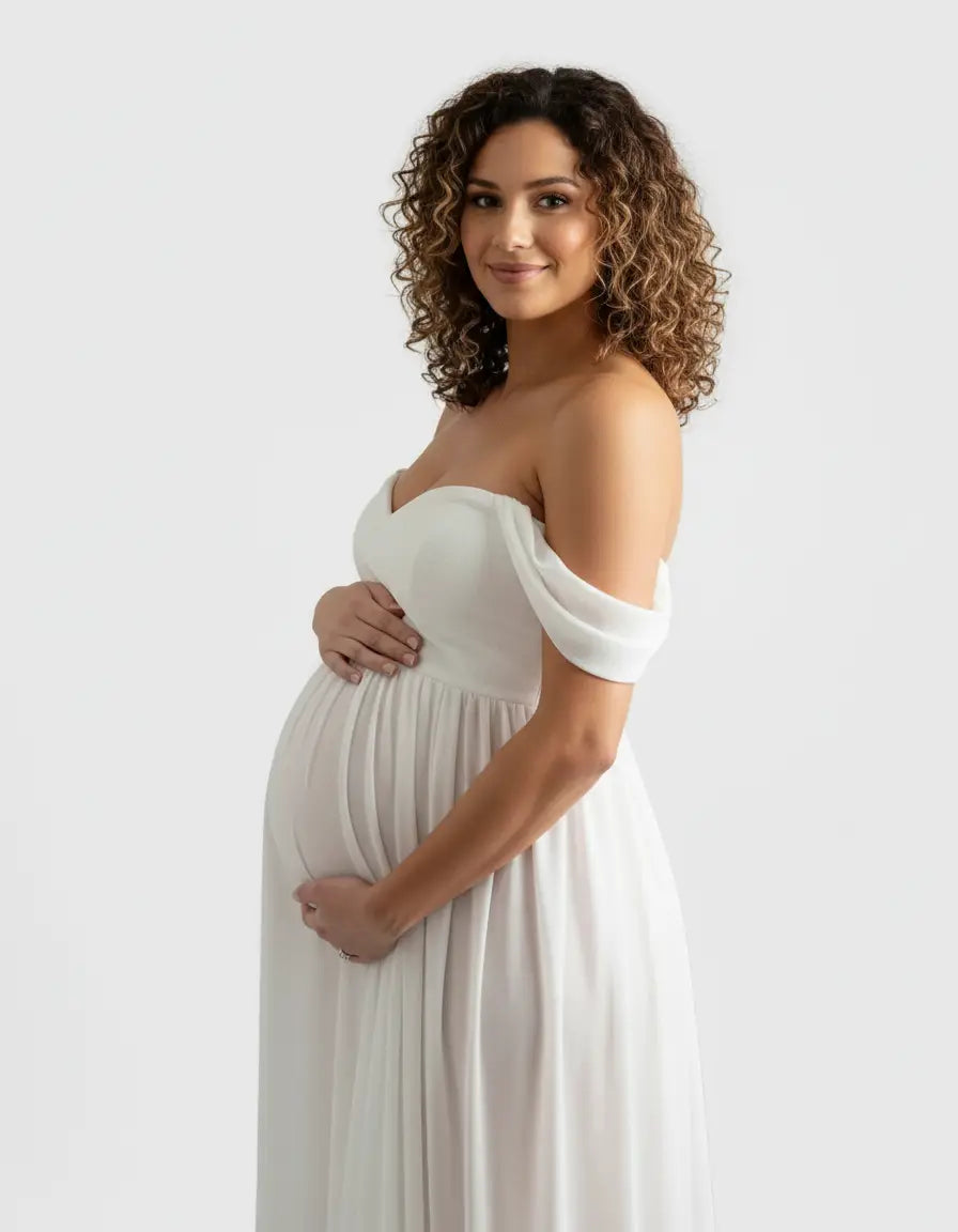 Smiling pregnant woman in a white off-shoulder flowing maternity photoshoot gown, cradling her bump.