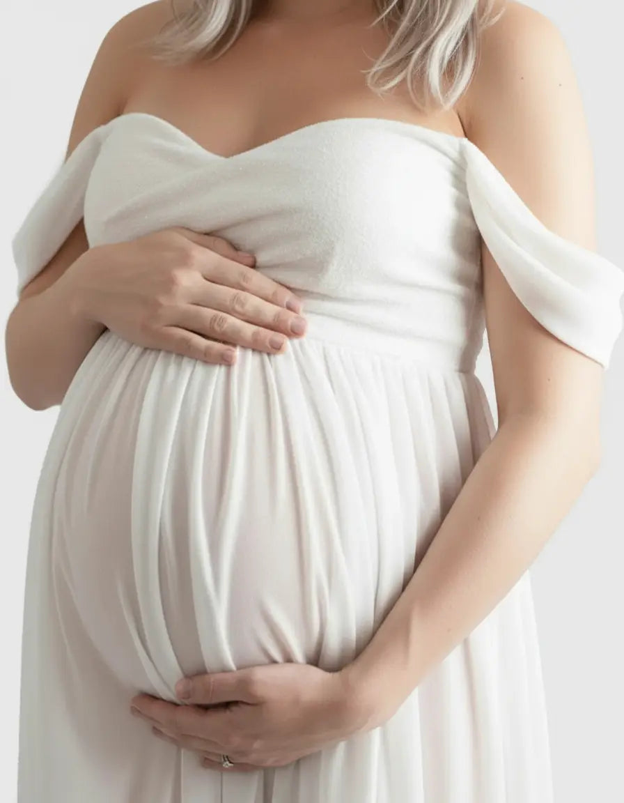 Pregnant woman in a white off-shoulder flowing maternity photoshoot gown, cradling her baby bump.