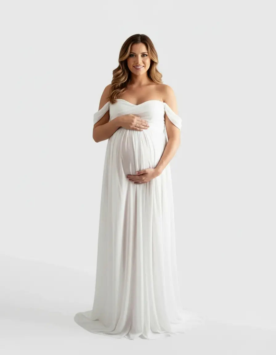 Smiling pregnant woman in a white off-shoulder flowing maternity photoshoot gown, hands on belly.