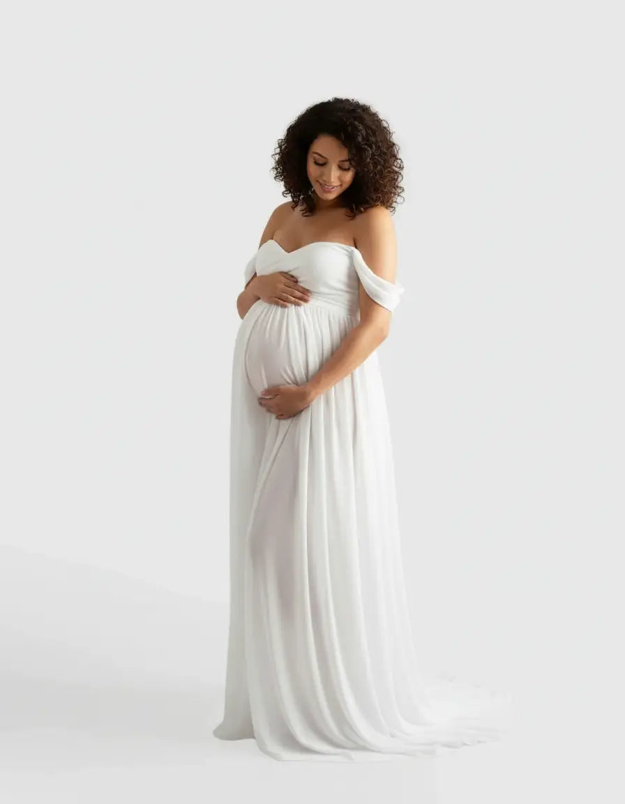 Pregnant woman in a white off-shoulder flowing maternity photoshoot gown, cradling her baby bump.