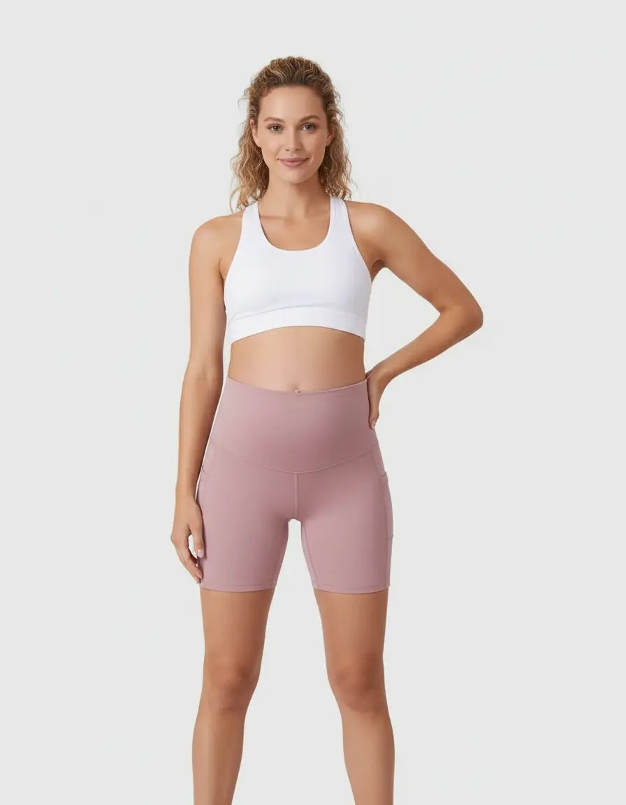 Smiling pregnant woman in pink over-belly maternity bike shorts with pockets and a white sports bra.