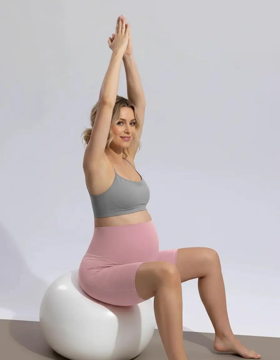 Pregnant woman in pink over-belly maternity bike shorts and grey bra, sitting on a white exercise ball.