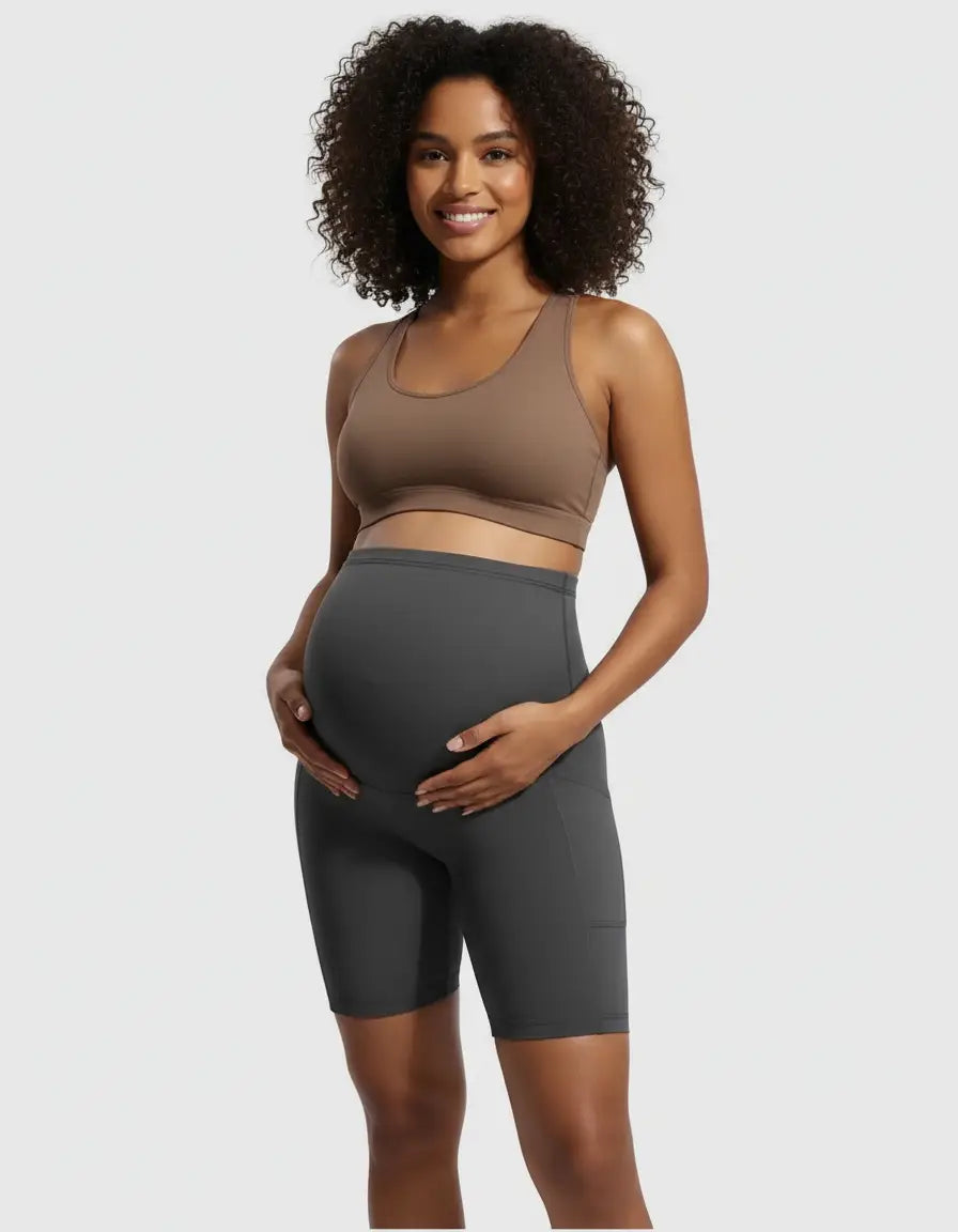 Smiling pregnant woman in charcoal over-belly maternity bike shorts with pockets and a brown sports bra.