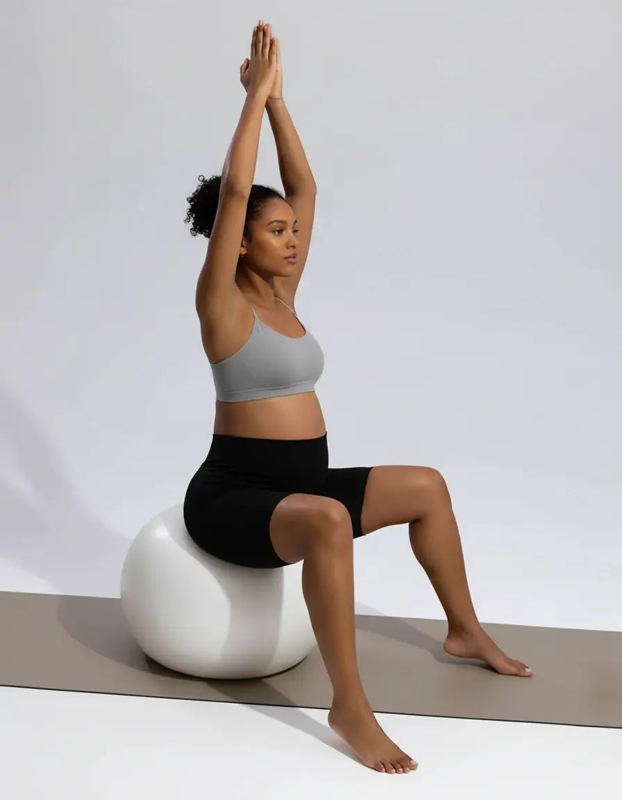 Pregnant woman in gray bra and black over-belly maternity bike shorts with pockets, sitting on a white exercise ball.
