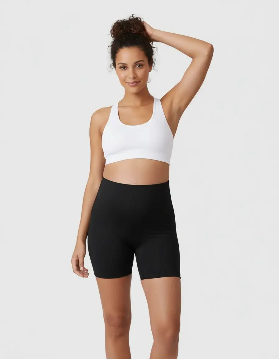 Smiling pregnant woman models black over-belly maternity bike shorts with pockets and a white bra.