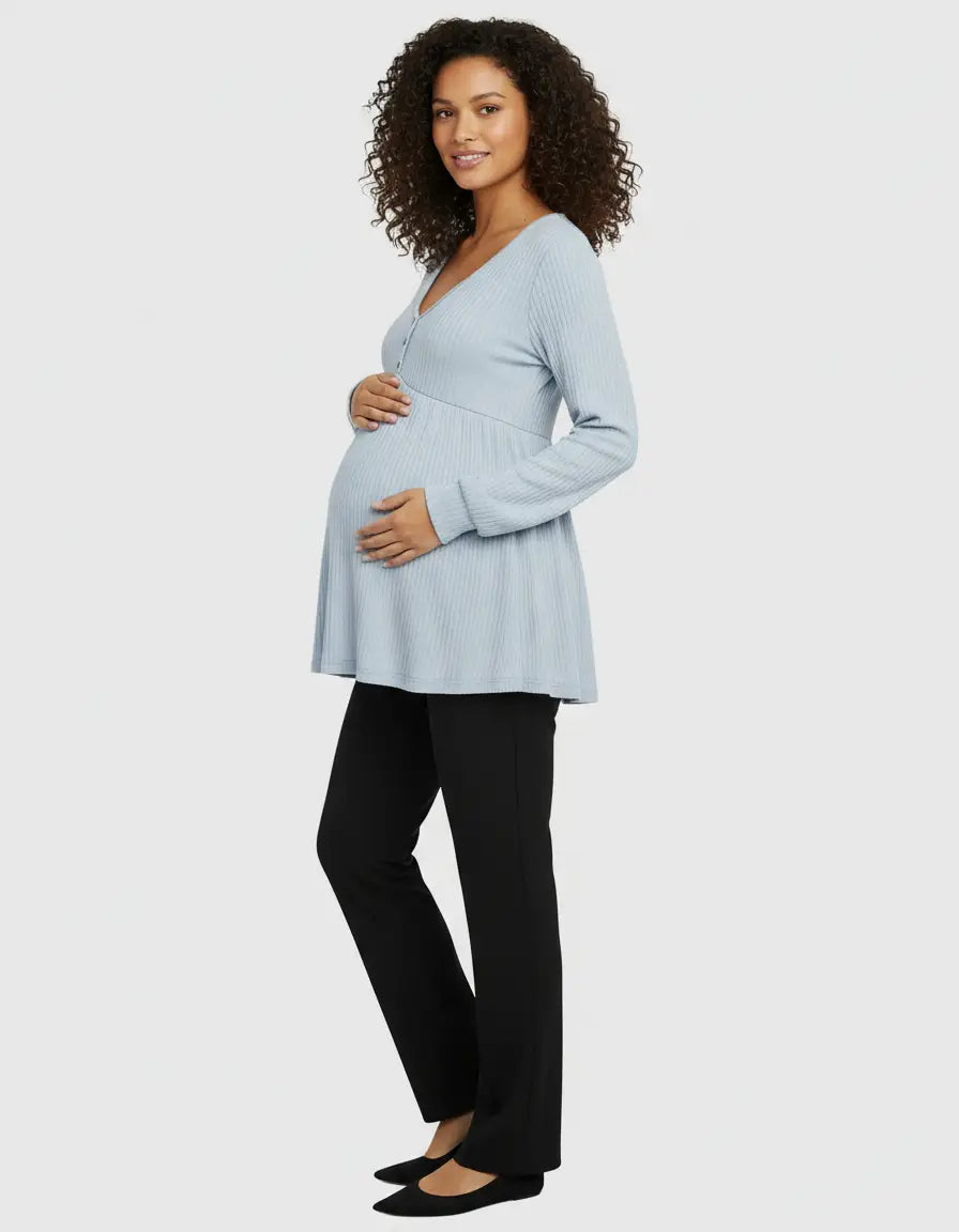 Smiling pregnant woman in a light blue ribbed V-neck maternity top with ruffled hem and black pants.