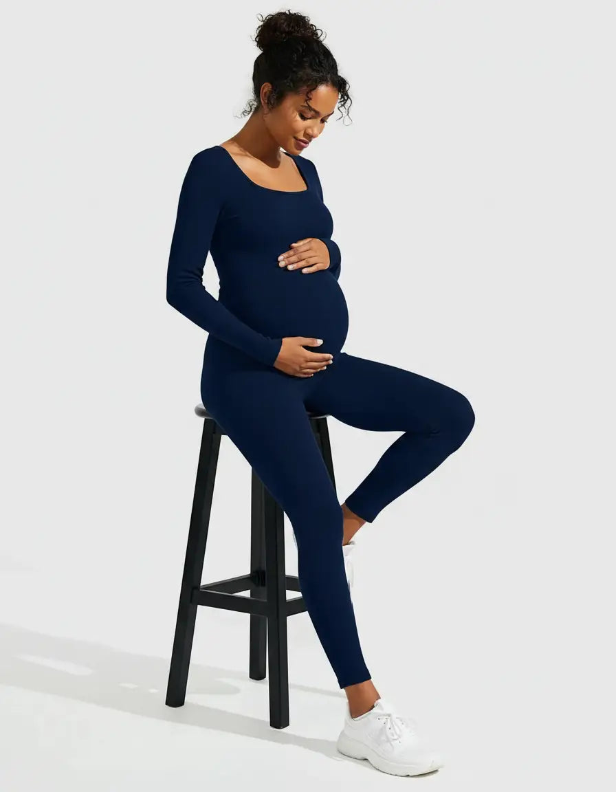 Pregnant woman in a navy blue seamless ankle-length maternity bodysuit, long sleeves, square neck, sitting on a stool.