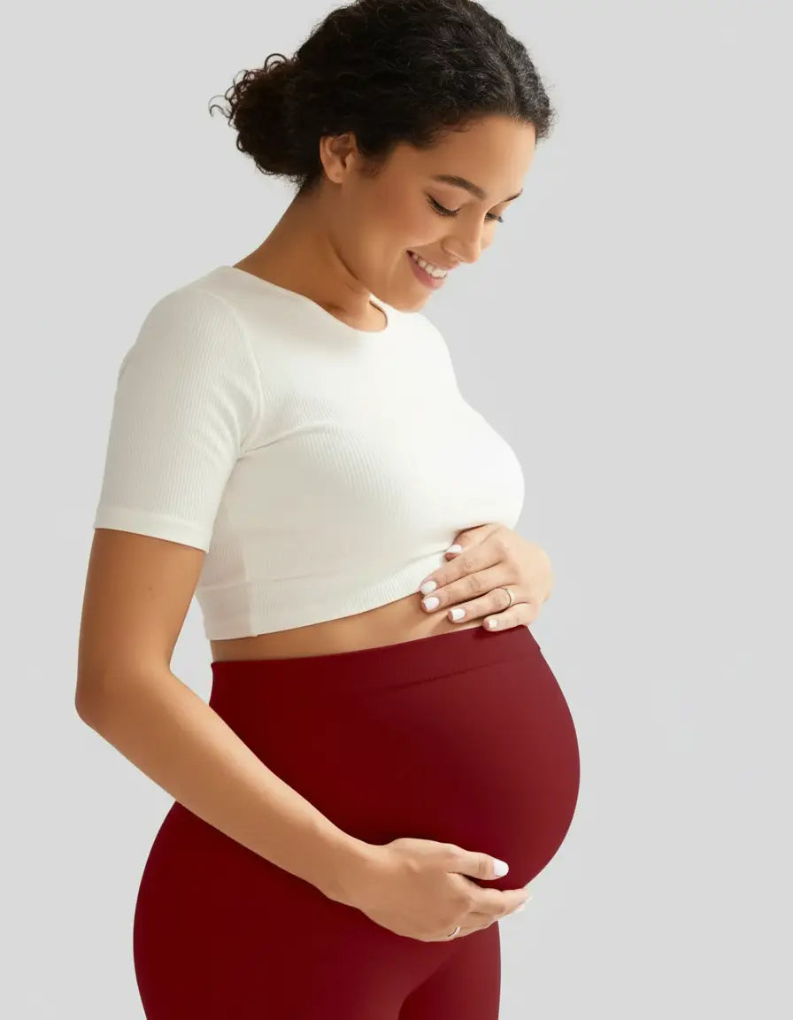 Smiling pregnant woman in a white ribbed crop top and dark red soft-stretch full-length maternity leggings.