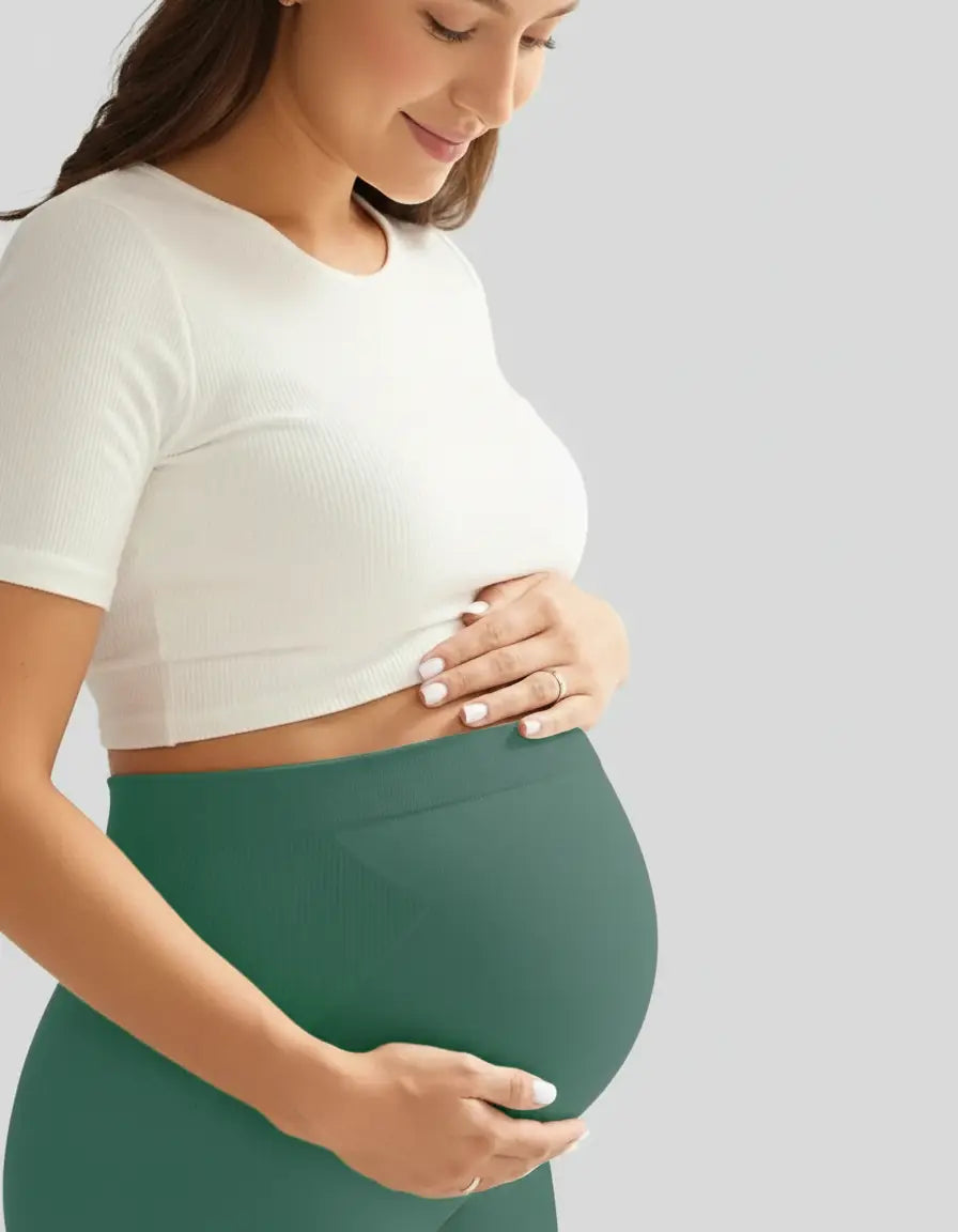 Smiling pregnant woman in a white ribbed crop top and soft-stretch full-length forest green maternity leggings.
