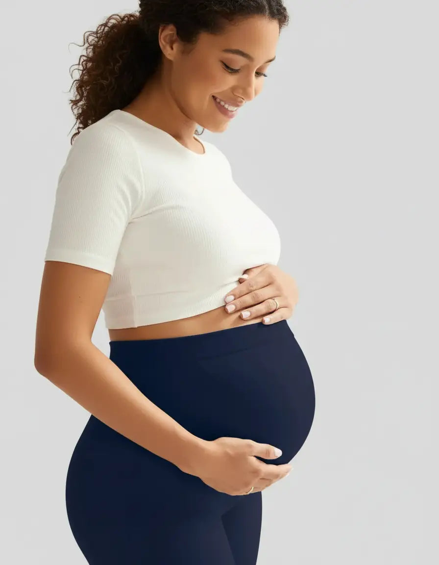 Smiling pregnant woman in soft-stretch navy full-length maternity leggings and a white crop top, cradling her bump.
